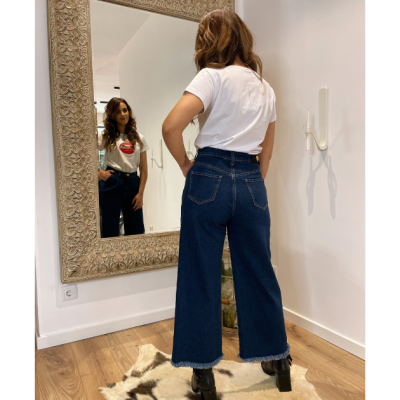 Mulher de costas usando calças largas de ganga azul escuro e t-shirt branca, olhando para um espelho grande com moldura decorativa