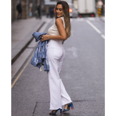 Mulher na rua com calças brancas largas, blusa branca e casaco azul no braço