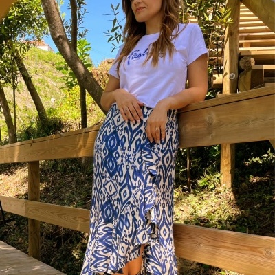 Mulher com camisola branca 'Flashy', saia azul e branca estampada e botins prateados à beira de um corrimão de madeira ao ar livre