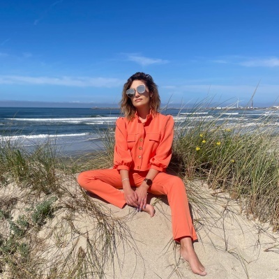 Mulher sentada na areia com conjunto de roupa laranja e óculos de sol espelhados junto ao mar