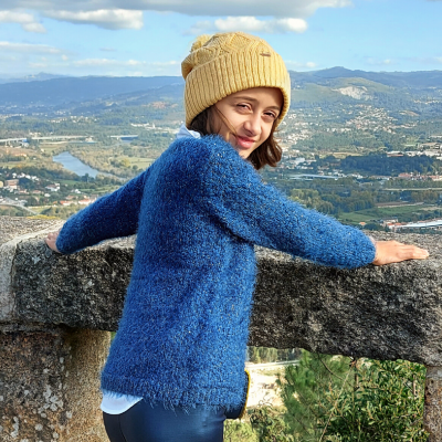 Mulher com suéter azul felpudo e gorro amarelo posando junto a guarda-corpo de pedra com paisagem ao fundo.