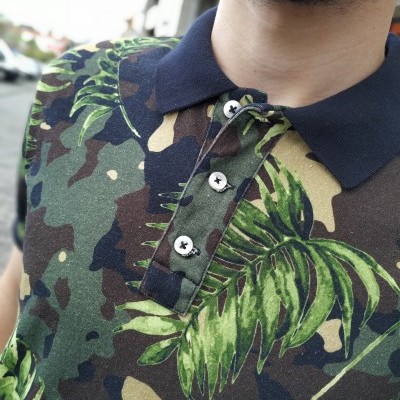 Polo com padrão militar e folhas tropicais verdes, gola azul escuro, botões brancos, em ambiente exterior
