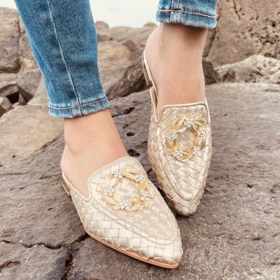 Mules dourados com padrão trançado e detalhe floral usados em ambiente rochoso