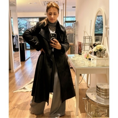 Pessoa com casaco preto longo, camisola cinzenta e calças largas, numa loja com chão de madeira e móveis brancos