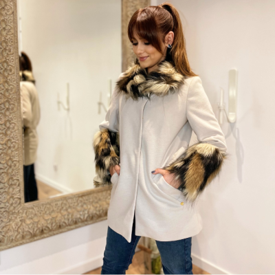 Casaco bege com gola e punhos em pelo sintético castanho e preto, mulher com cabelo castanho, interior com espelho e parede branca