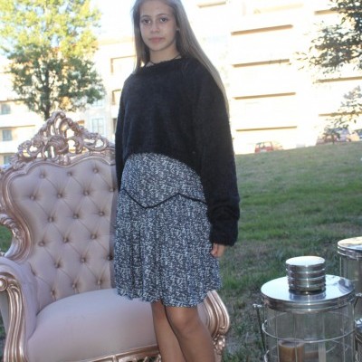 Mulher jovem com vestido cinza claro com padrão e camisola preta em pé ao lado de cadeira rosa estofada e mesa metálica com velas.