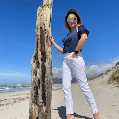 Mulher na praia com calças brancas e t-shirt azul ao lado de um tronco de madeira