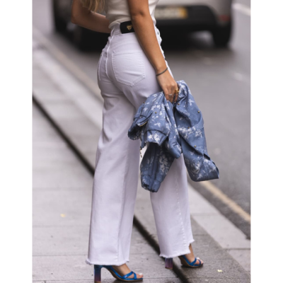 Calças brancas de ganga de perna larga com casaco azul tie-dye na mão e sandálias azuis numa rua