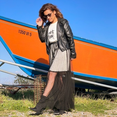 Mulher com casaco preto de pele e saia longa preta com transparências ao lado de barco laranja e azul