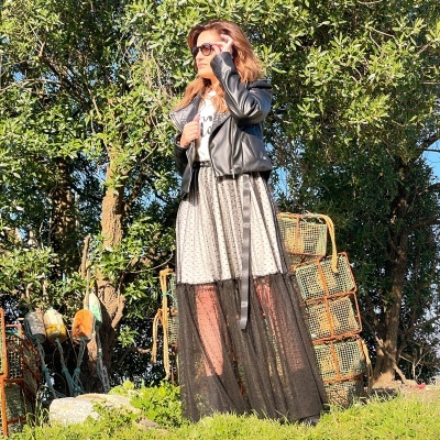 Mulher com vestido longo preto e branco, jaqueta de pele preta e óculos de sol em ambiente ao ar livre