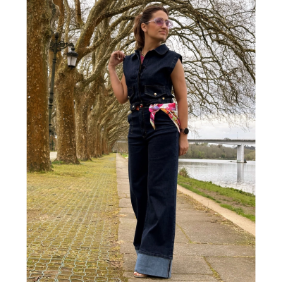 Mulher com macacão de ganga preto com cinto colorido, ao ar livre junto a rio e árvores.