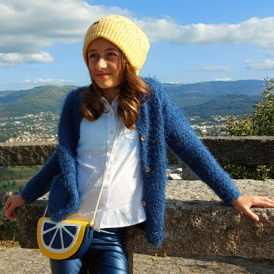 Menina a usar casaco azul, gola branca, gorro e carteira amarelos, encostada a muro de pedra