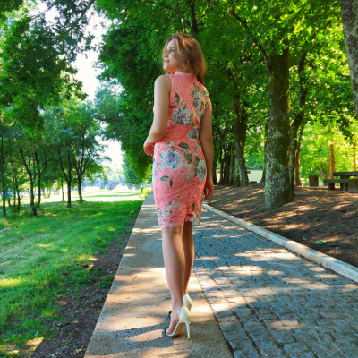 Mulher de costas a usar vestido cor de salmão com padrão floral e saltos altos, em passeio arborizado.