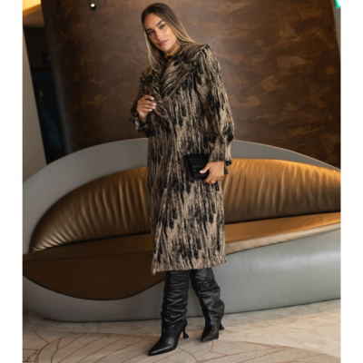 Mulher vestindo casaco comprido estampado castanho e preto, botas pretas e carteira preta, em ambiente interno com sofá cinza e castanho.