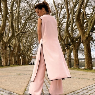 Mulher de costas com colete longo rosa claro e calças largas, na rua com árvores despidas