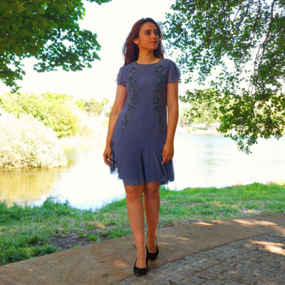 Mulher a usar vestido azul curto com aplicações florais verdes, em ambiente exterior junto a lago