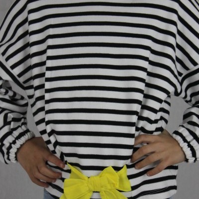 Camisola branca com riscas pretas e laço amarelo, criança de mãos na cintura.