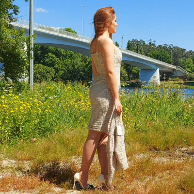 Mulher de vestido bege de malha junto a rio e ponte ao fundo