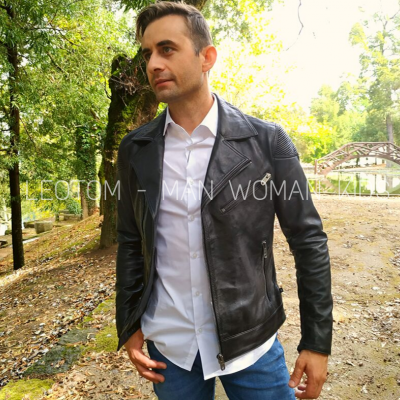 Homem veste jaqueta de couro preta, camisa branca e calças azuis, ao ar livre num parque com árvores e ponte