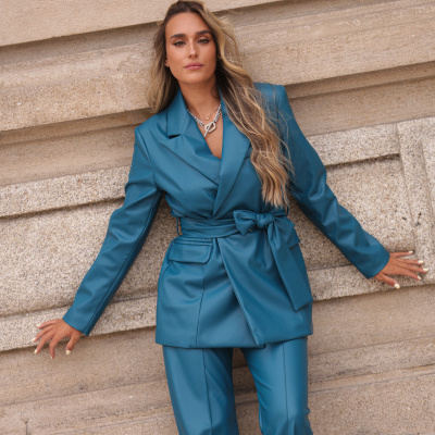 Fato azul acetinado com blazer com laço e calças compridas, mulher encostada numa parede de pedra clara.