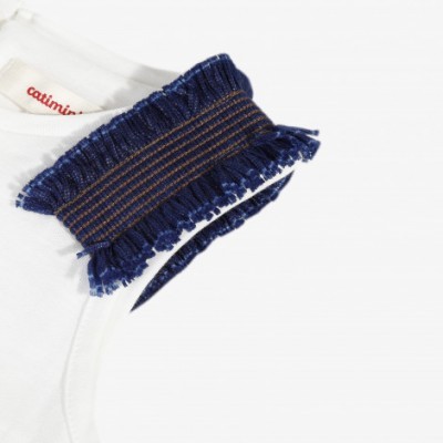 Detalhe de ombro de roupa branca com franjas azul e etiqueta com texto vermelho