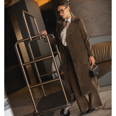 Mulher com casaco castanho de veludo e camisa branca ao lado de carrinho de bagagem metálico