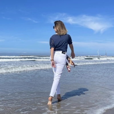 Mulher na praia com t-shirt azul, calças brancas, lenço colorido e óculos escuros