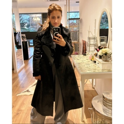 Casaco preto com botões e gola de pelo, pessoa a tirar selfie num espelho, sala com chão de madeira, mesa branca com tampo de vidro e espelho circular na parede