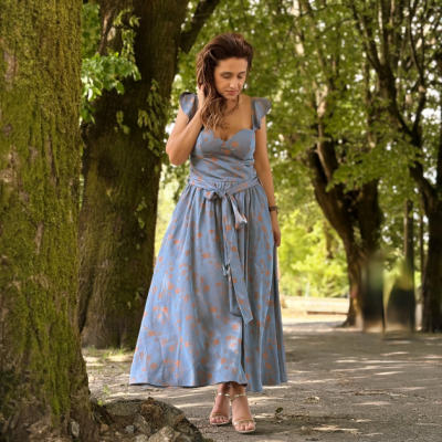 Mulher com vestido comprido azul com padrão floral laranja, posando ao ar livre entre árvores