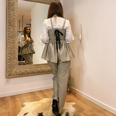 Conjunto feminino com blusa branca, colete cinzento riscado com laço preto nas costas e calças riscas, em cenário interior com espelho e tapete de pele.