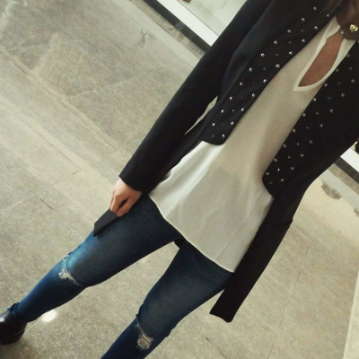 Pessoa vestindo blazer preto com pérolas, camisa branca, jeans azul escuro rasgados e sapatos pretos, chão de pedra e prateleira ao fundo