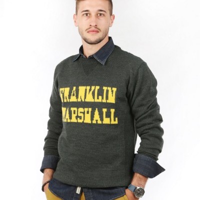 Homem com camisola de malha verde com texto amarelo, camisa azul e calças amarelas em fundo branco.