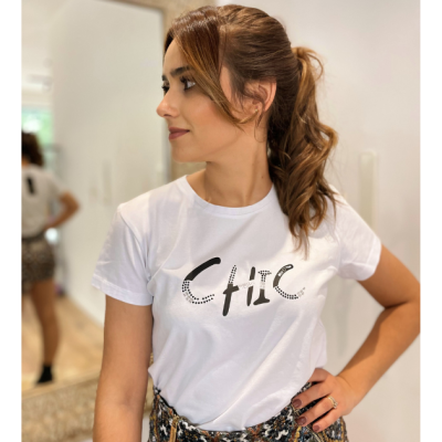 Mulher a usar t-shirt branca com estampado 'Chic', cabelo castanho apanhado, ambiente interior com paredes brancas e espelho