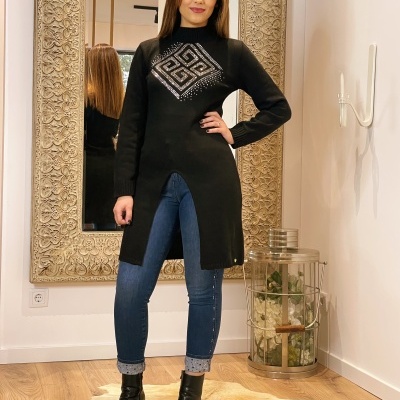 Mulher com camisola preta comprida com padrão geométrico branco, calças de ganga e botas pretas em divisão interior com espelho grande e chão de madeira