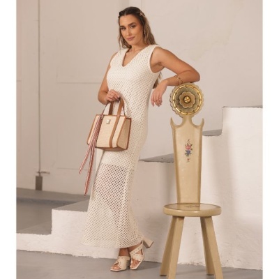 Mulher com vestido branco de malha segurando mala bege junto a cadeira decorativa