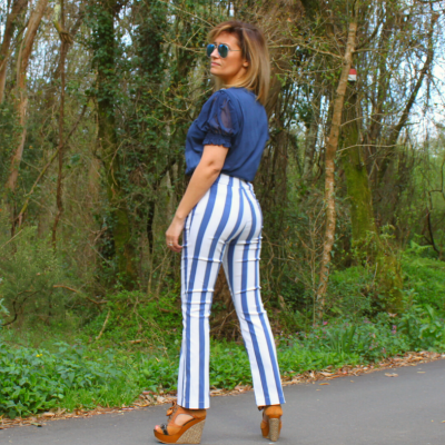 Mulher de costas vestindo calças listradas brancas e azuis, blusa azul e sapatos altos castanhos numa estrada florestada