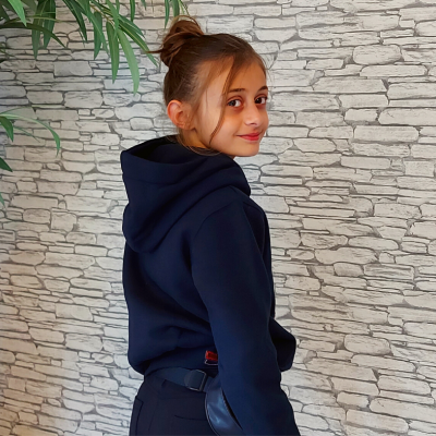 Menina de perfil com capuz preto e parede de pedra ao fundo