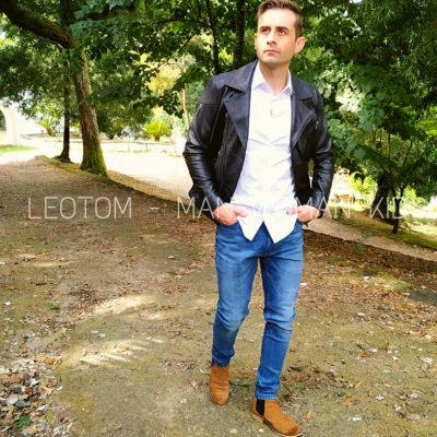 Homem com camisa branca, blazer preto e calças de ganga azuis a caminhar num parque com árvores