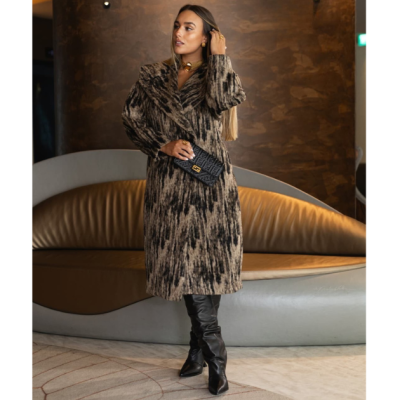 Mulher com casaco longo castanho e preto, botas altas pretas e carteira preta em ambiente interior