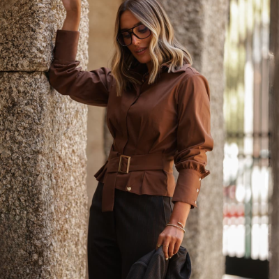 Mulher com roupa castanha e calças escuras junto a uma parede de pedra