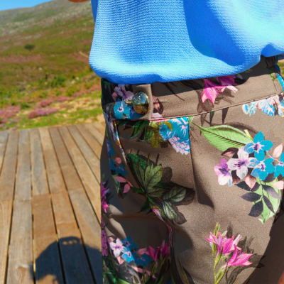Calças florais castanhas com botão grande, camisa azul à cintura, ambiente exterior com plataforma de madeira.