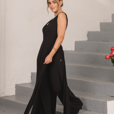 Mulher com vestido preto comprido e fluido ao lado de escadas cinza, parede branca e flores vermelhas.