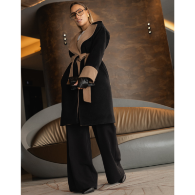 Mulher com casaco preto e bege, calças pretas largas e óculos, em sala com sofá cinza e castanho