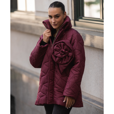 Casaco matelassado bordeaux com flor decorativa e fecho dourado, usado por mulher junto a parede urbana