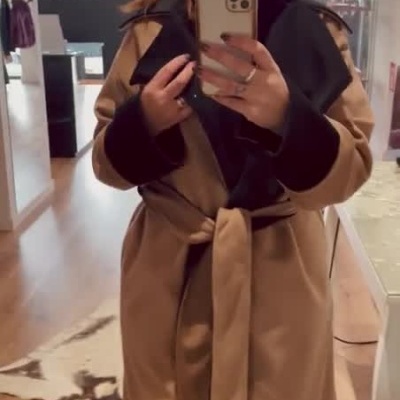 Mulher a experimentar casaco bege com detalhes pretos e cinto, tirando foto num espelho