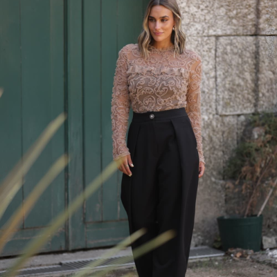 Mulher com blusa transparente nude com padrão e calças pretas largas em ambiente exterior