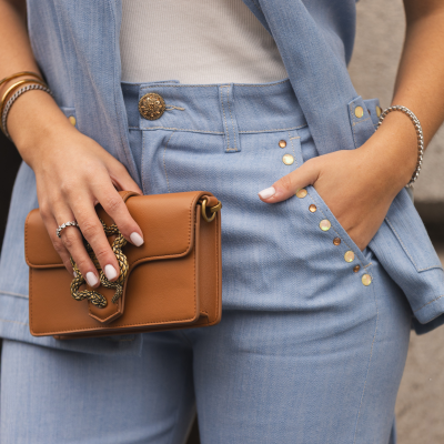 Conjunto de colete e calças de ganga azul claro com botões dourados, top branco, mala castanha com fivela metálica, anéis e pulseiras