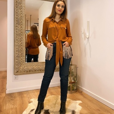 Pessoa com camisa castanha com detalhes prateados, calças pretas e botas, em quarto com espelho e tapete de pele