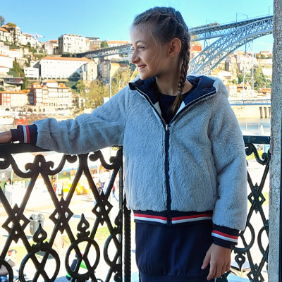 Criança com casaco felpudo cinzento claro em varanda com vista de cidade e ponte
