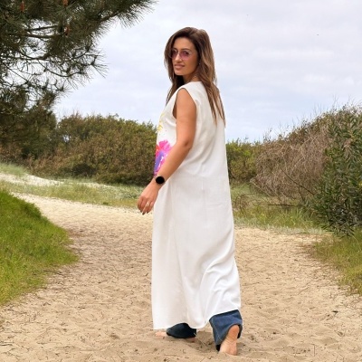 Mulher com vestido branco e calça azul em caminho de areia com vegetação